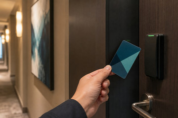 A hand presenting a plastic hotel key card to a contactless reader beside a hotel room door